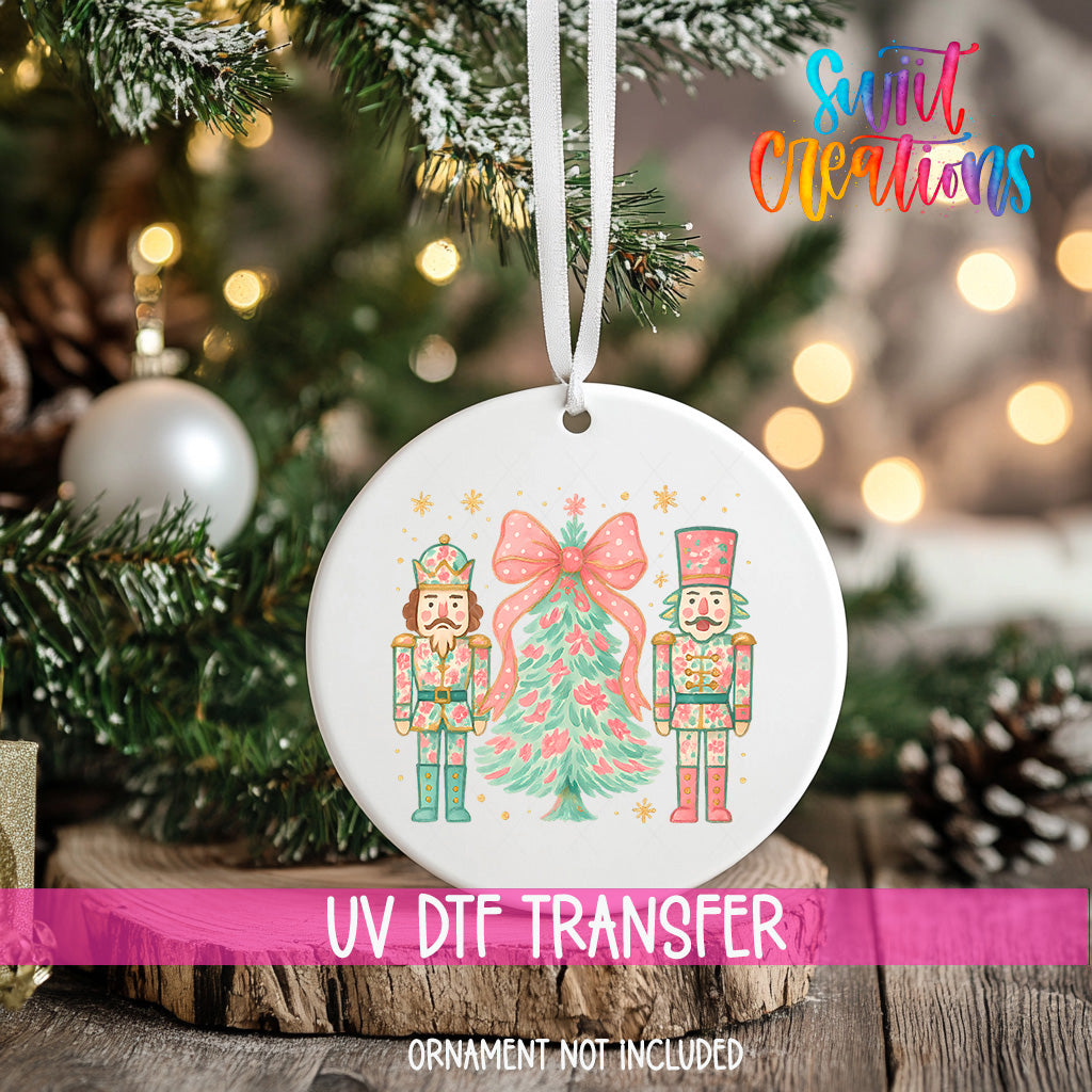 A white ornament with a Christmas tree design and two nutcracker characters is hanging on a Christmas tree.