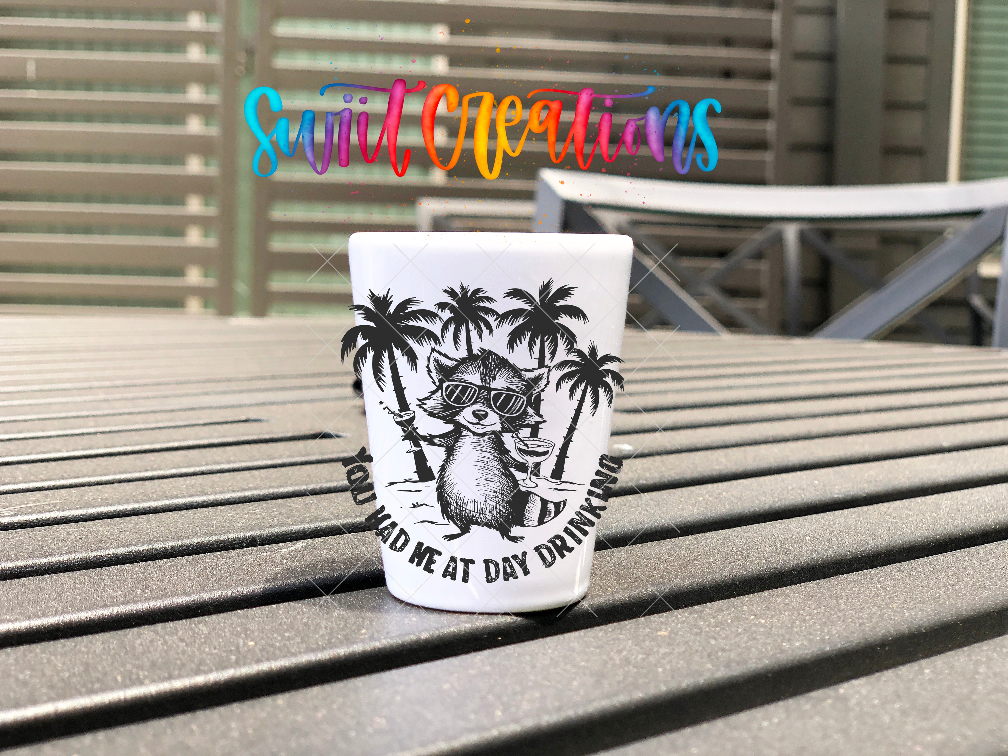 A white shot glass with a black and white illustration of a raccoon wearing sunglasses and holding a drink, sitting on a wooden surface.