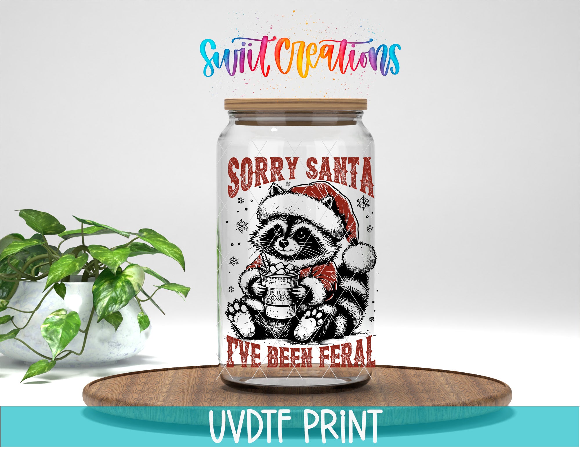 A glass jar with a raccoon wearing a Santa hat and holding a cup, with the text "Sorry Santa I've been feral!" printed on it. The jar is placed on a wooden surface, and there is a potted plant in the background.