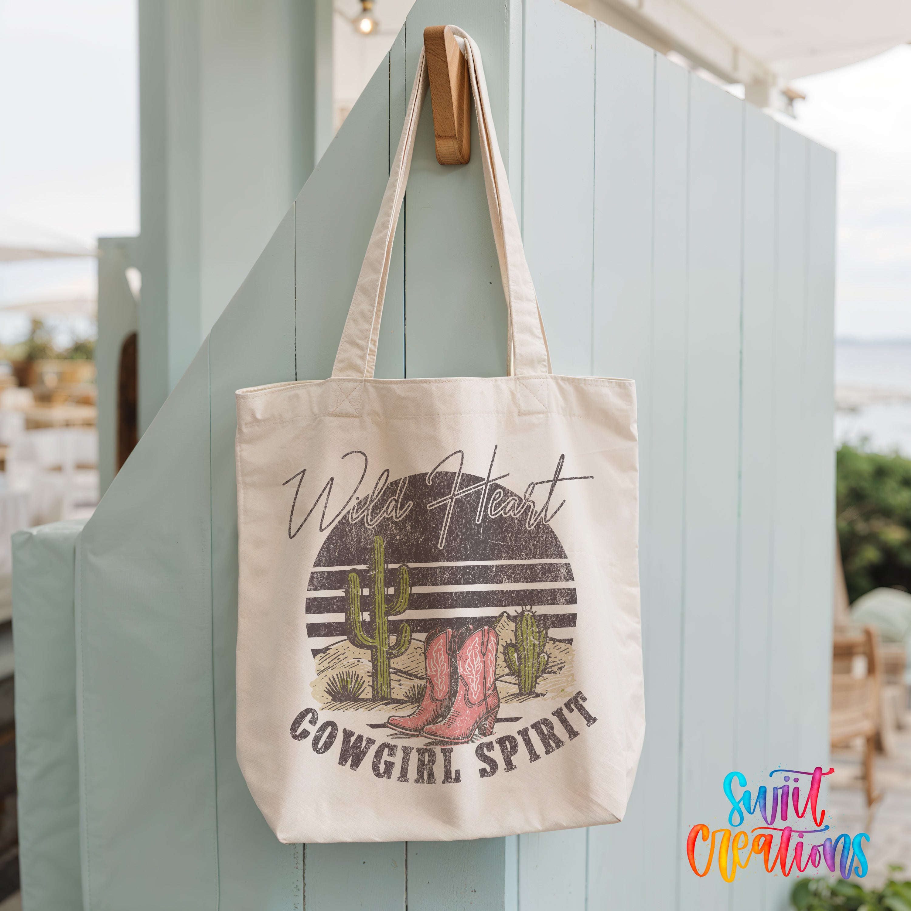 A white tote bag with a graphic design of a cactus and cowboy boots, along with the text "Wild Heart Cowgirl Spirit" printed on it.