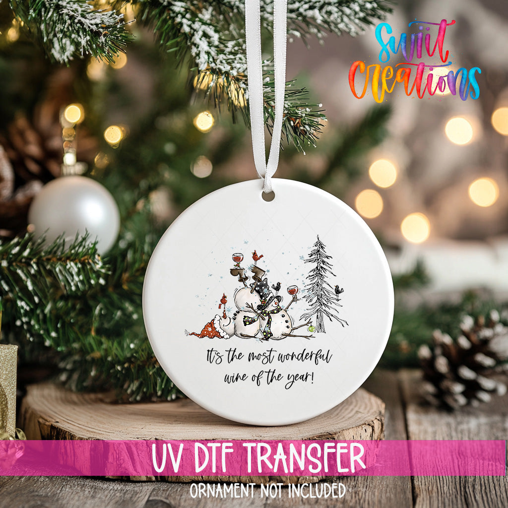 A white ceramic ornament with a snowman and pine trees on it, hanging on a Christmas tree.