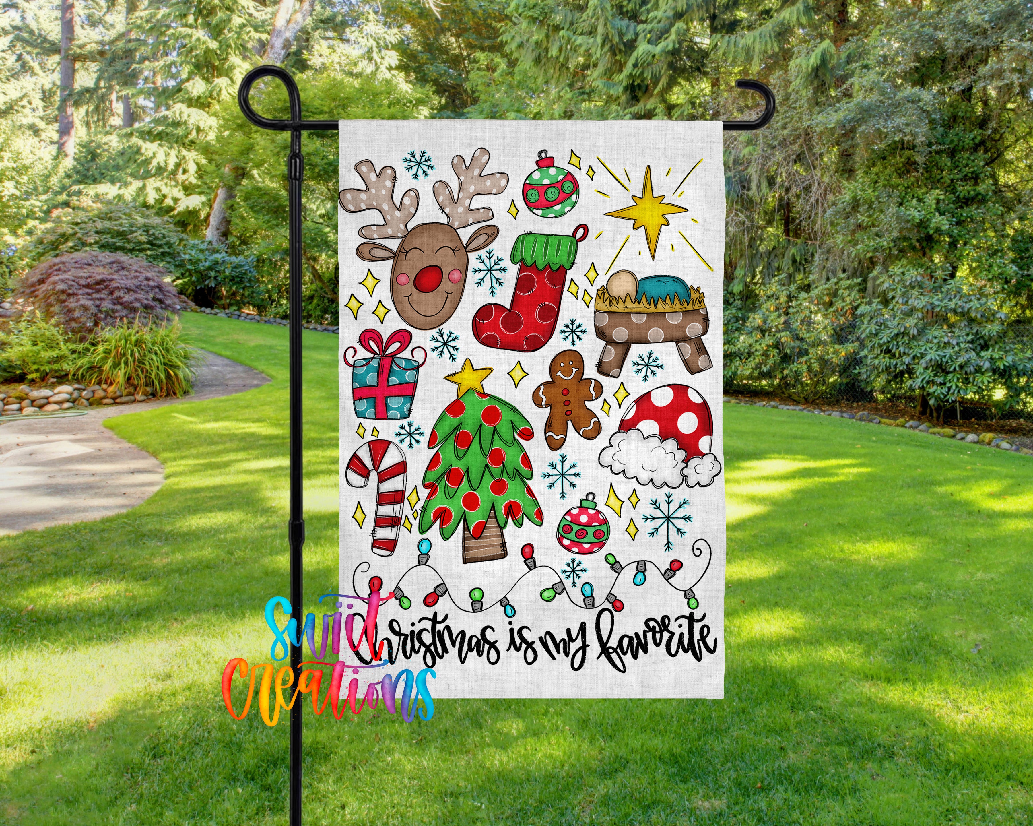 A colorful Christmas-themed flag is displayed in a garden setting, featuring various festive elements such as reindeer, a nativity scene, a star, and other holiday decorations.