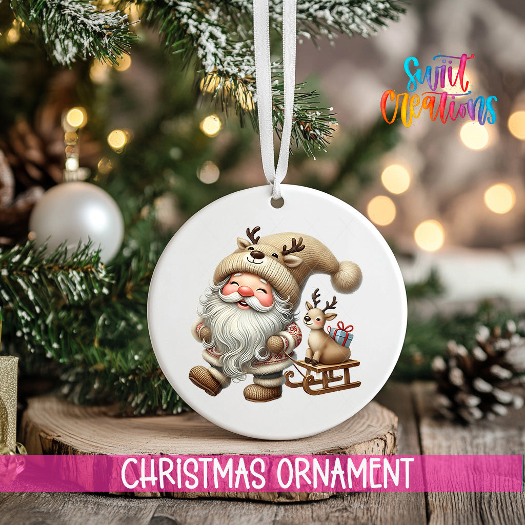 A white Christmas ornament featuring a smiling Santa Claus with reindeer and a sled, hanging on a tree branch with a blurred background of lights and pine cones.