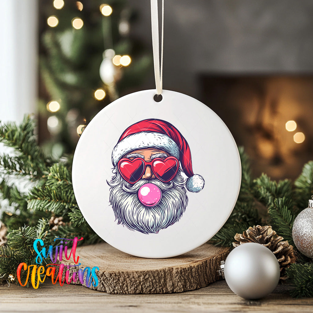 A white ornament with a Santa Claus face wearing sunglasses and a red hat, with a pink bubble gum bubble in his mouth, is displayed on a wooden surface.