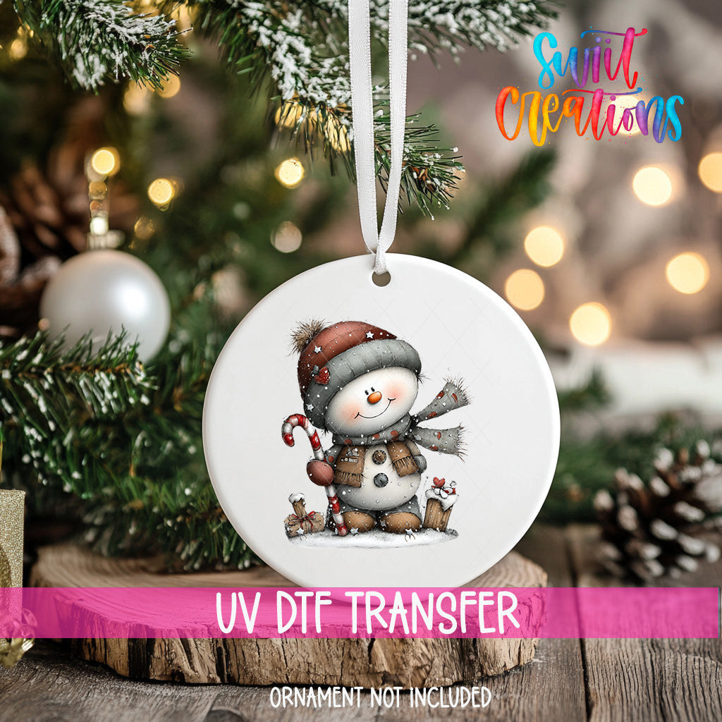 A white ornament with a snowman design hangs on a Christmas tree, surrounded by pine branches and twinkling lights.