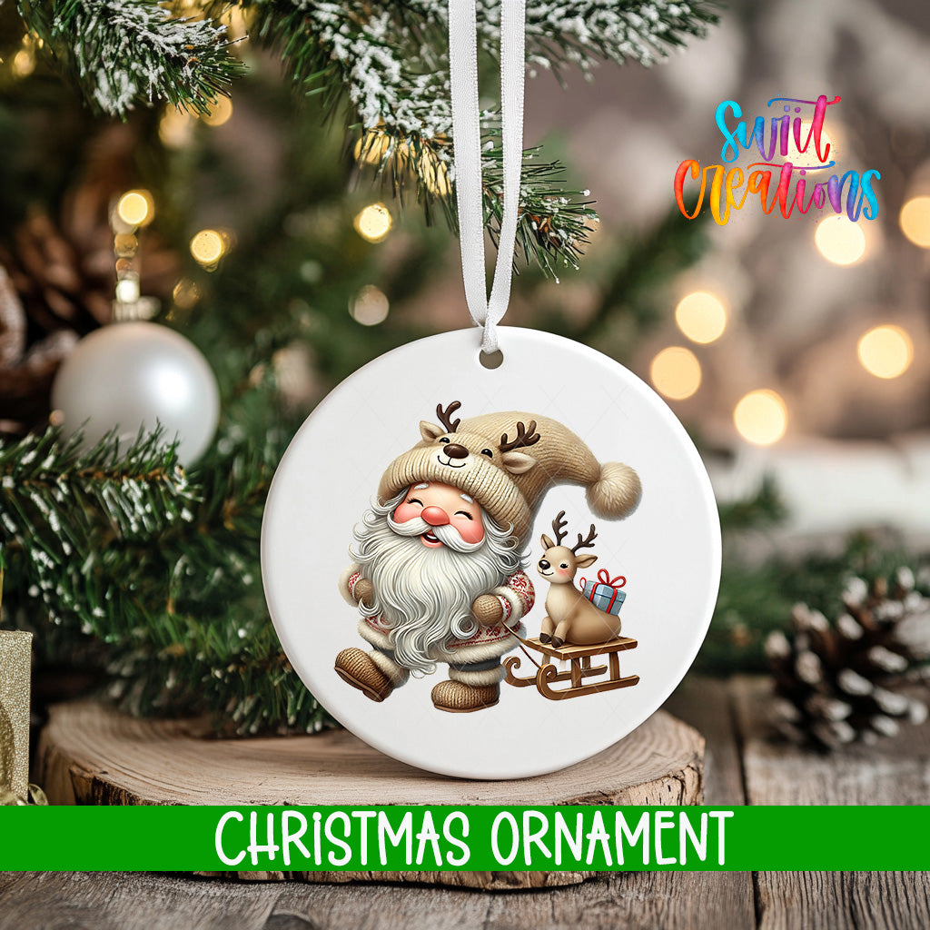 A white Christmas ornament featuring a Santa Claus figure with reindeer and a sled, hanging on a tree branch with a blurred background of lights.