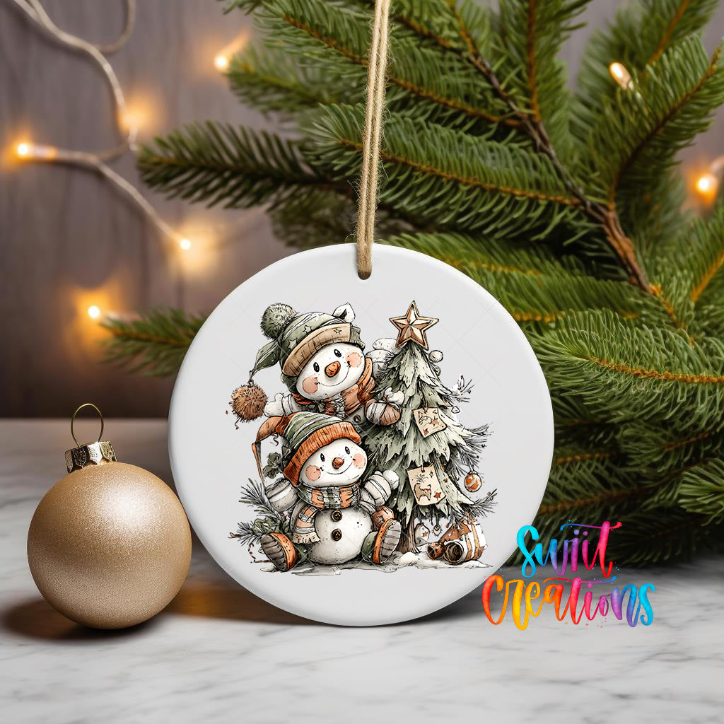 A white ceramic ornament featuring two snowmen sitting under a Christmas tree, surrounded by pine branches and other holiday decorations, with a gold ornament and twinkling lights in the background.