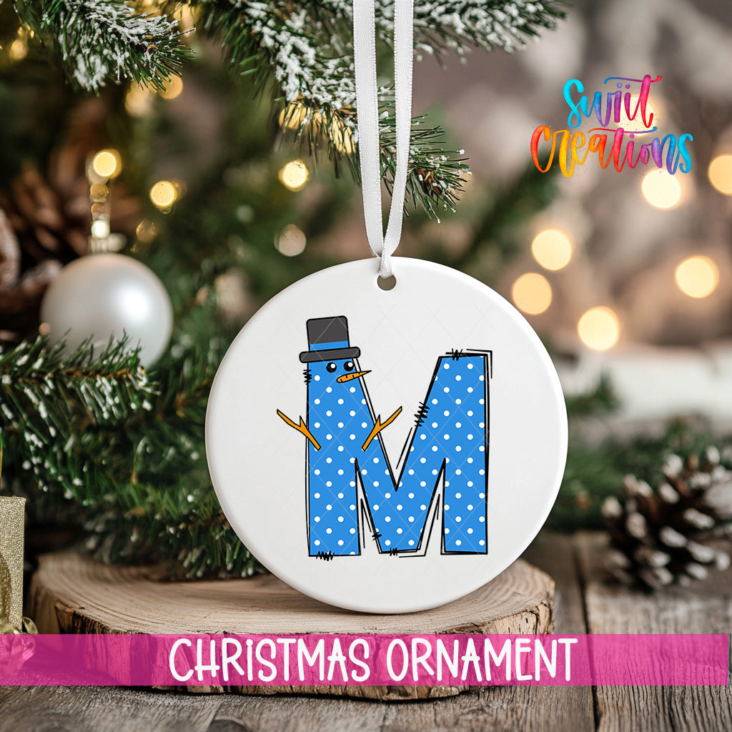 A Christmas ornament featuring a snowman made out of the letter "M" with blue polka dots, hanging on a tree branch.