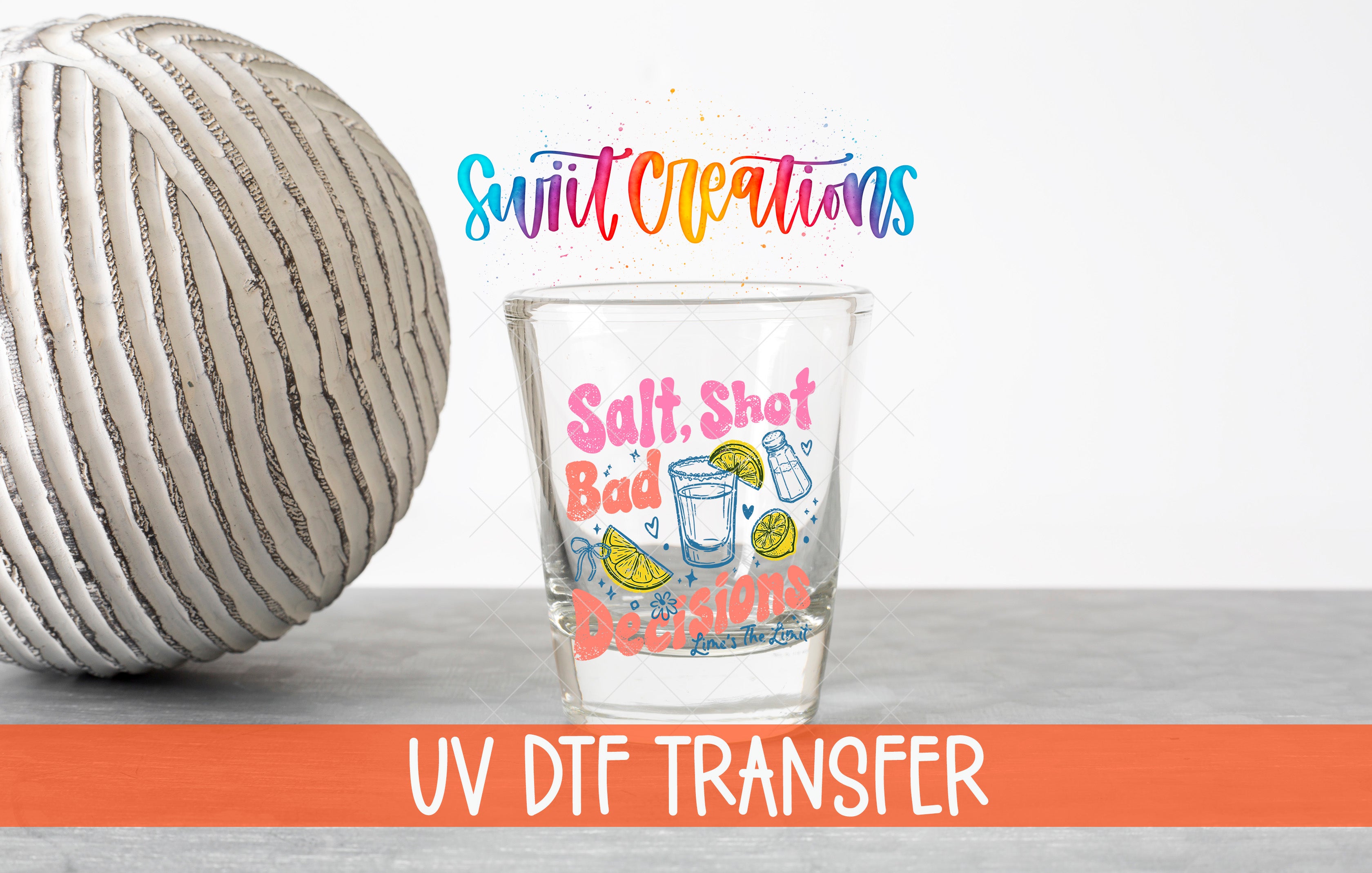 a shot glass with a colorful design and text, placed next to a gray ball on a white surface.