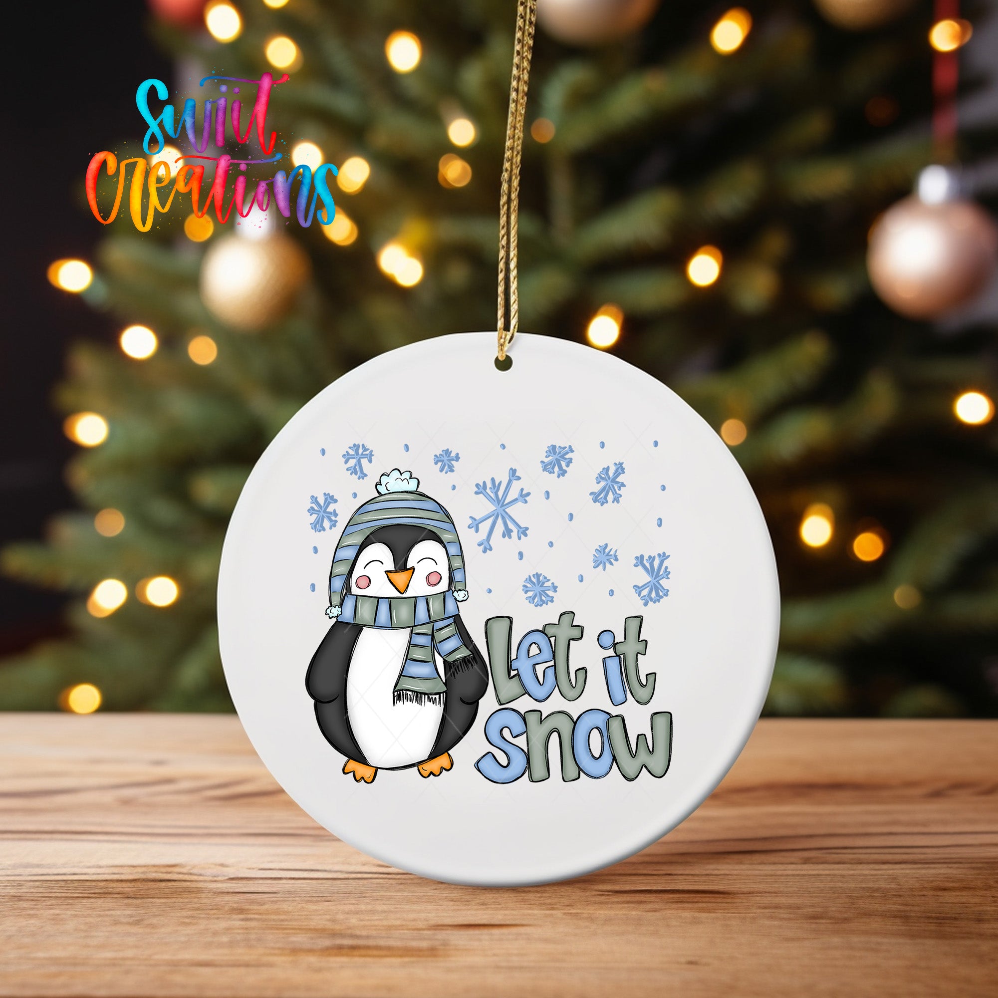 A white ornament with a cartoon penguin wearing a scarf and hat, along with the text "Let it Snow" and snowflakes, is hanging from a string.