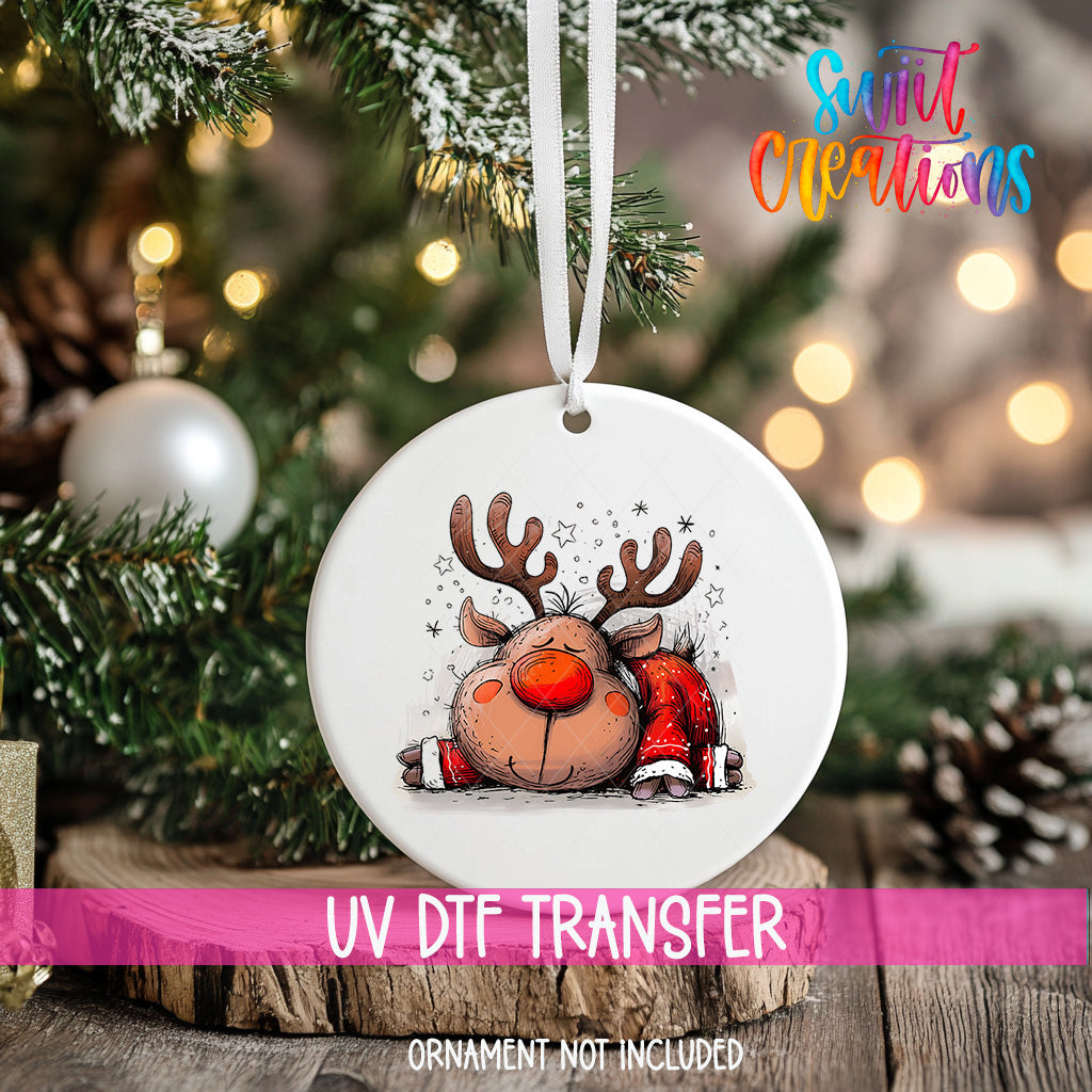 A white ornament with a cartoon reindeer design hangs on a Christmas tree, surrounded by pine branches and twinkling lights.