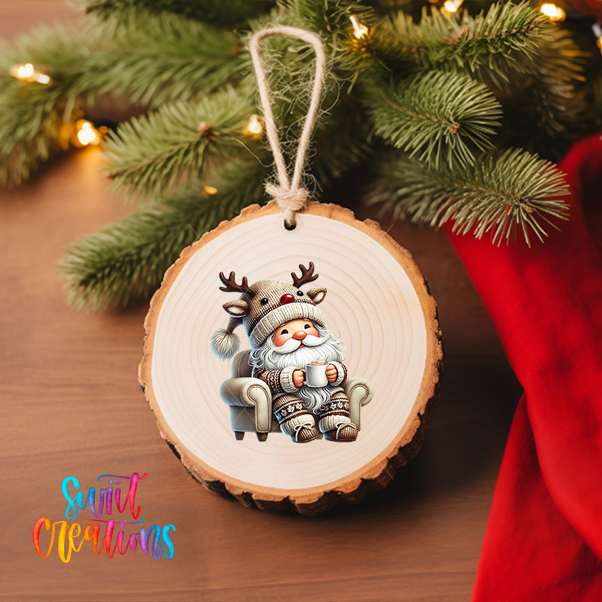 A wooden ornament featuring a reindeer sitting in a chair, with a Santa Claus-like figure wearing a hat and holding a cup of hot chocolate. The ornament is hanging from a Christmas tree, and there is a red cloth visible in the background.