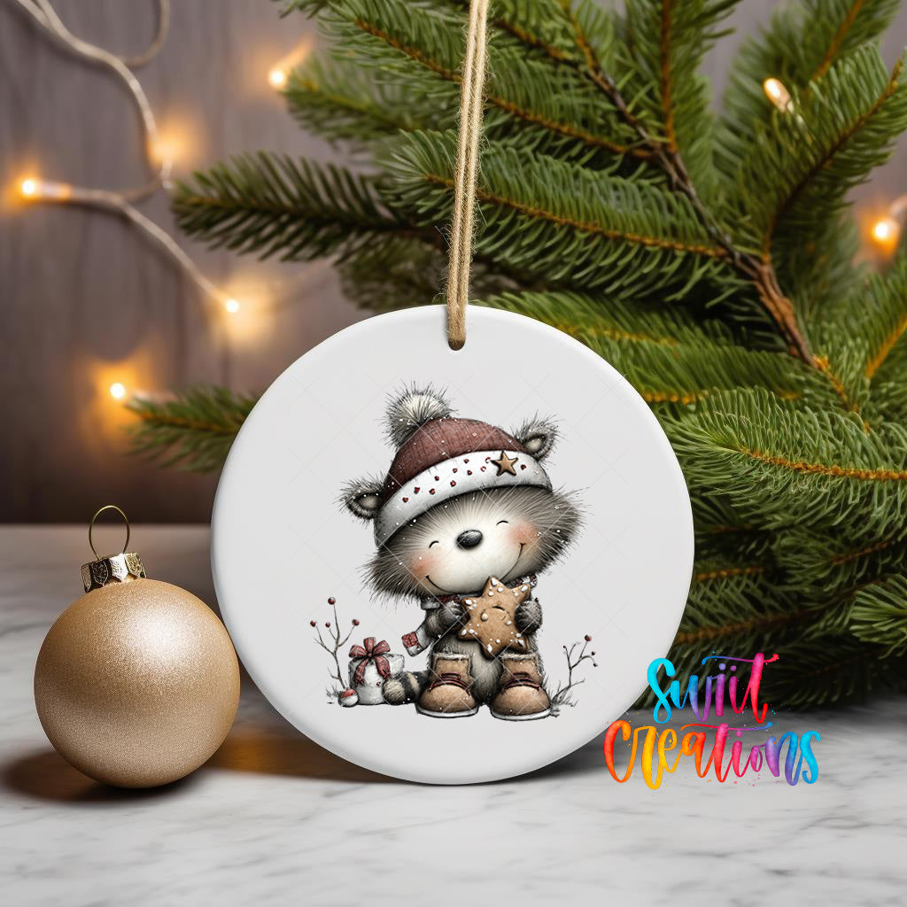 A white ceramic ornament featuring a cartoon image of a smiling bear wearing a hat and holding a star, placed on a marble surface next to a gold ornament and a Christmas tree with twinkling lights.
