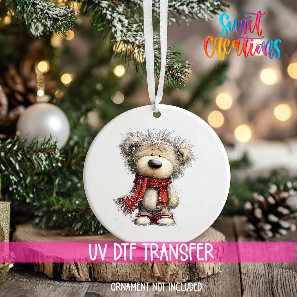 A white ornament with a teddy bear design hangs on a Christmas tree, surrounded by pine branches and twinkling lights.