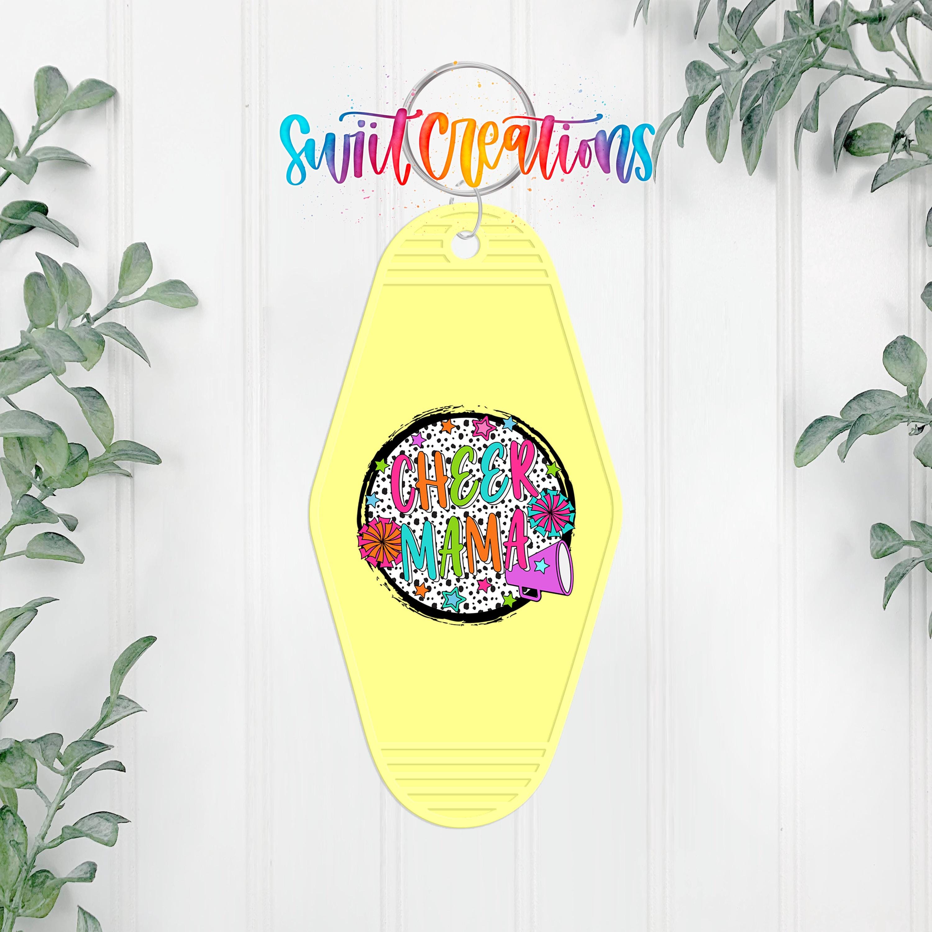 A yellow keychain with a colorful design and text, hanging on a white wall with greenery in the background.