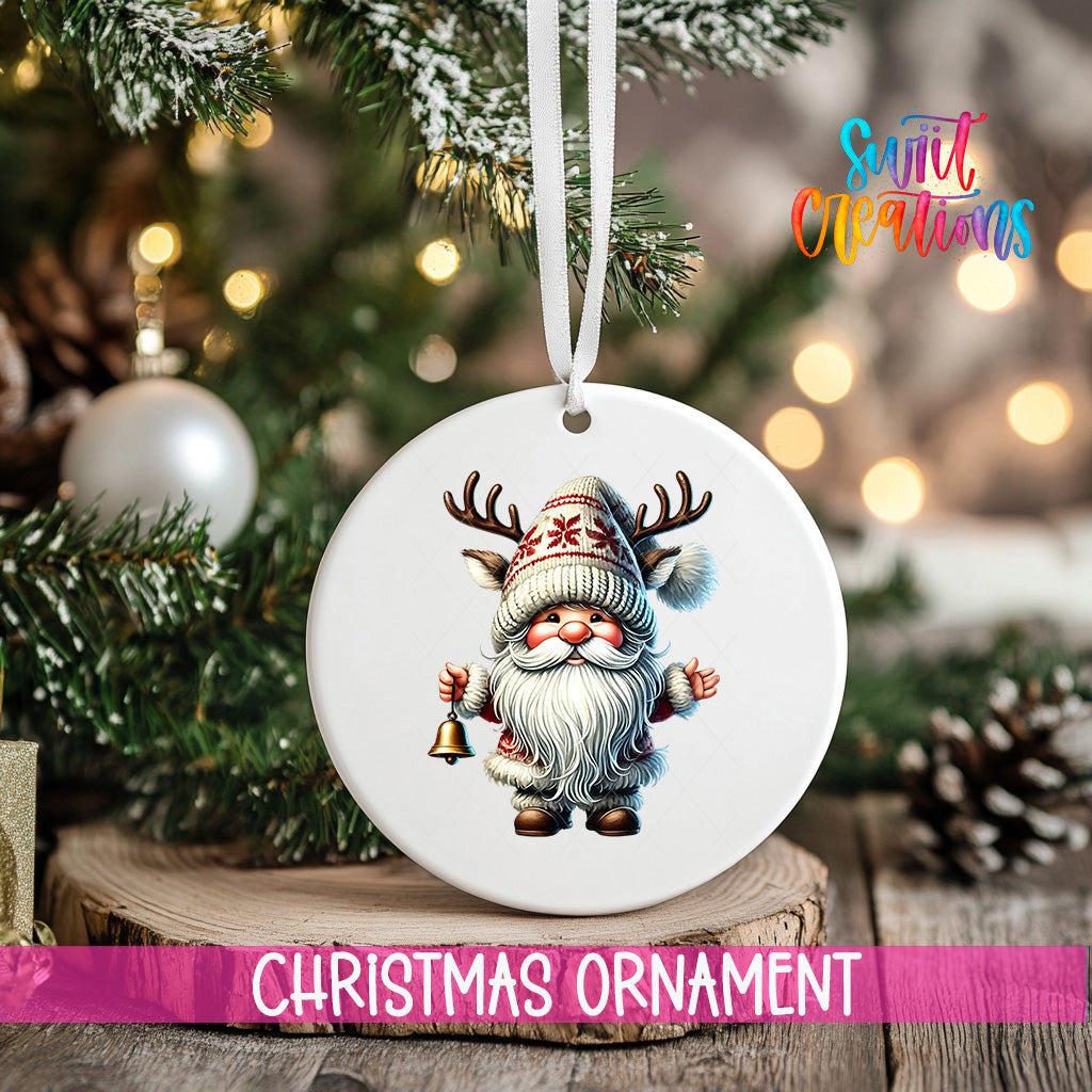 A white Christmas ornament featuring a gnome with antlers is hanging from a tree branch, with a blurred background of lights and pine cones.