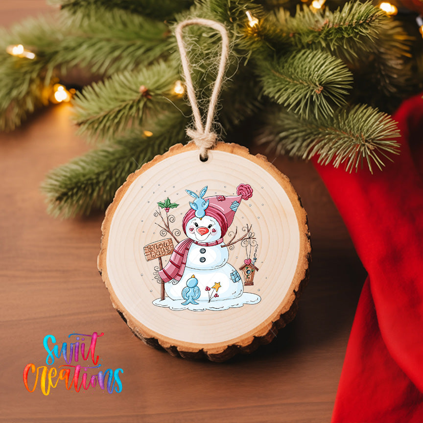 A wooden ornament featuring a snowman with a sign, surrounded by pine branches and twinkling lights, hangs on a Christmas tree.
