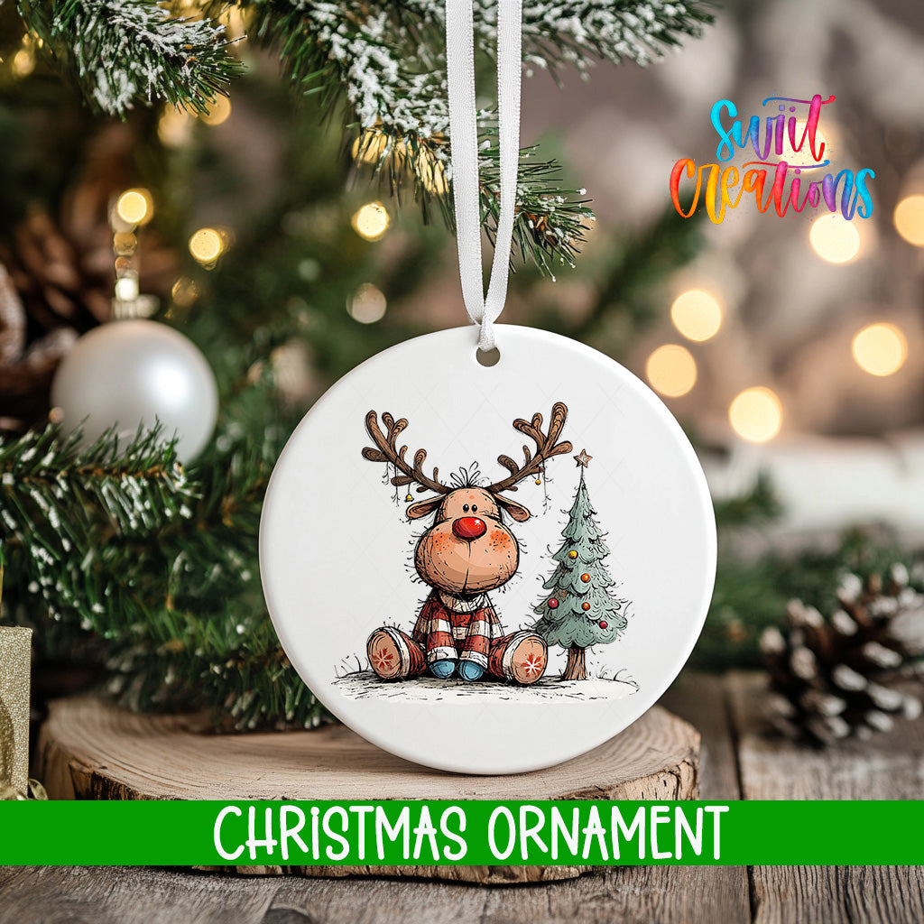A white Christmas ornament featuring a cartoon reindeer sitting next to a Christmas tree, with a blurred background of pine branches and twinkling lights.