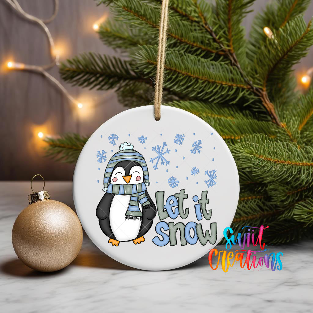 A white ornament with a cartoon penguin wearing a scarf and hat, along with the text "Let it snow" and snowflakes, is displayed on a table next to a gold ornament and a Christmas tree.