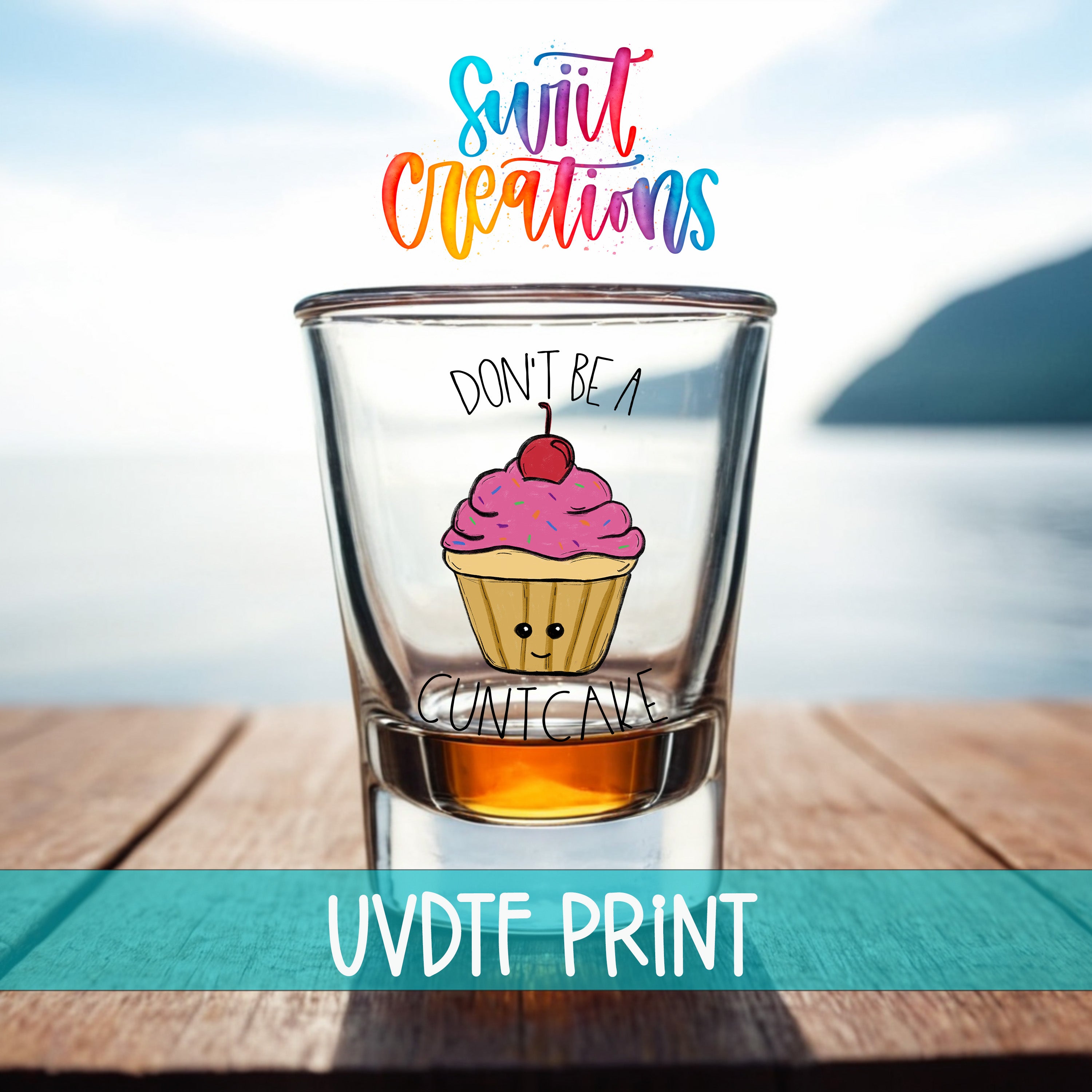 A shot glass with a cupcake design on it, placed on a wooden surface with a body of water in the background.
