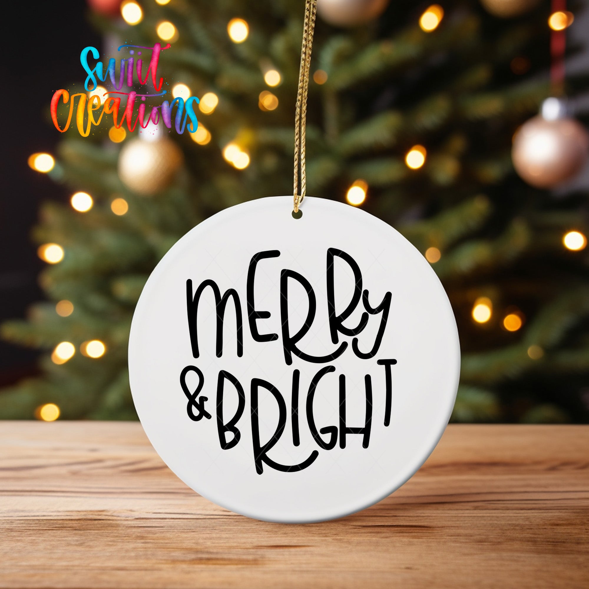 A white ceramic ornament with the text "MERRY & BRIGHT" written on it, hanging from a string. The ornament is placed on a wooden surface, with a blurred background featuring a Christmas tree and colorful lights.