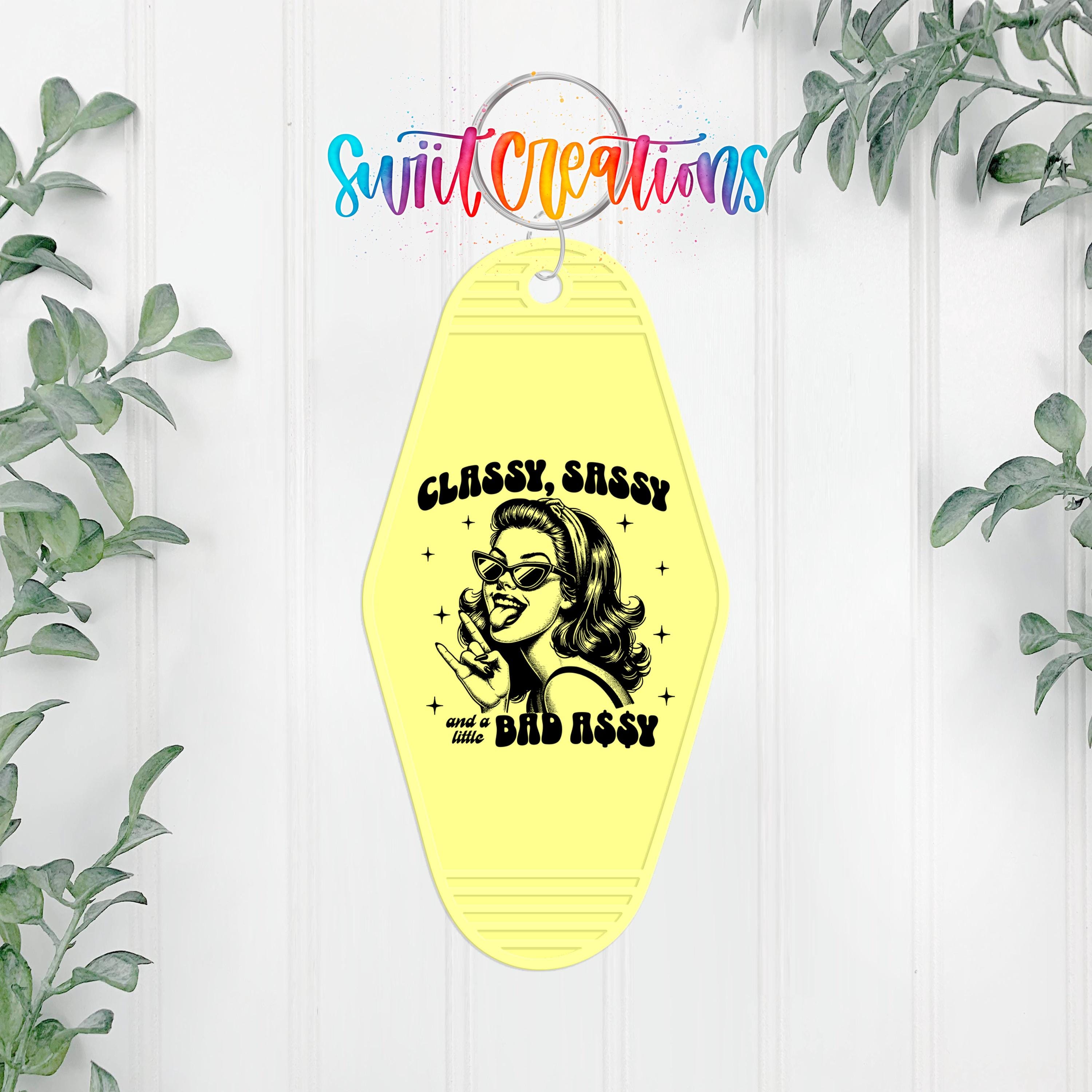 A yellow keychain with a black and white illustration of a woman and the text "CLASSY SASSY AND A LITTLE BAD ASSY" printed on it.