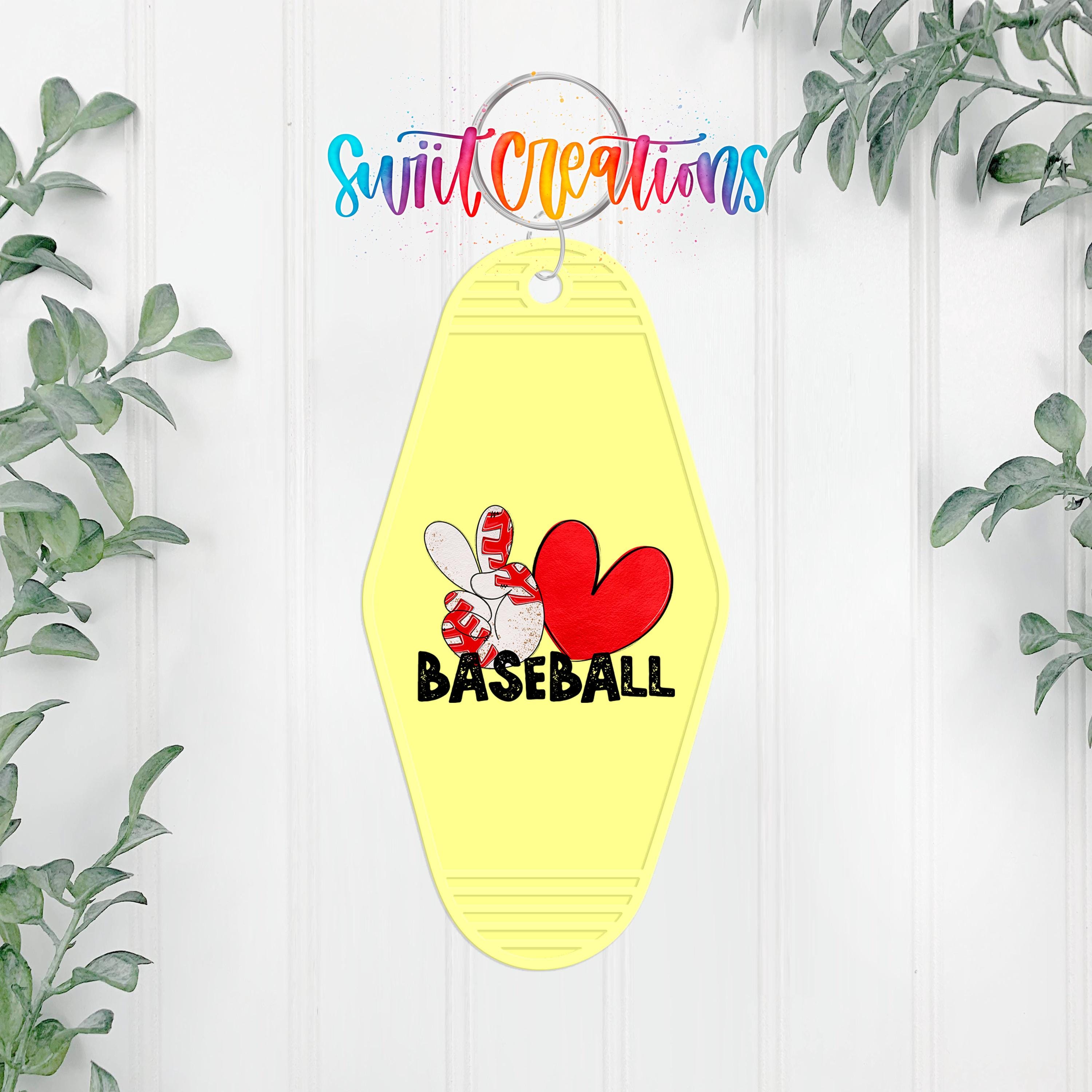 A yellow keychain with a red heart and the word "BASEBALL" printed on it, hanging on a white wall with greenery in the background.