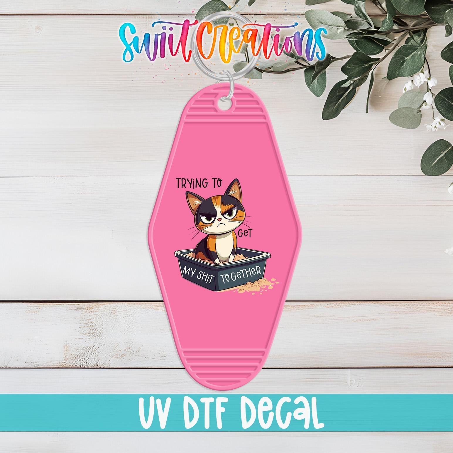 A pink keychain with a cartoon cat and the text "Trying to get my shit together" printed on it, placed on a wooden surface with some greenery in the background.