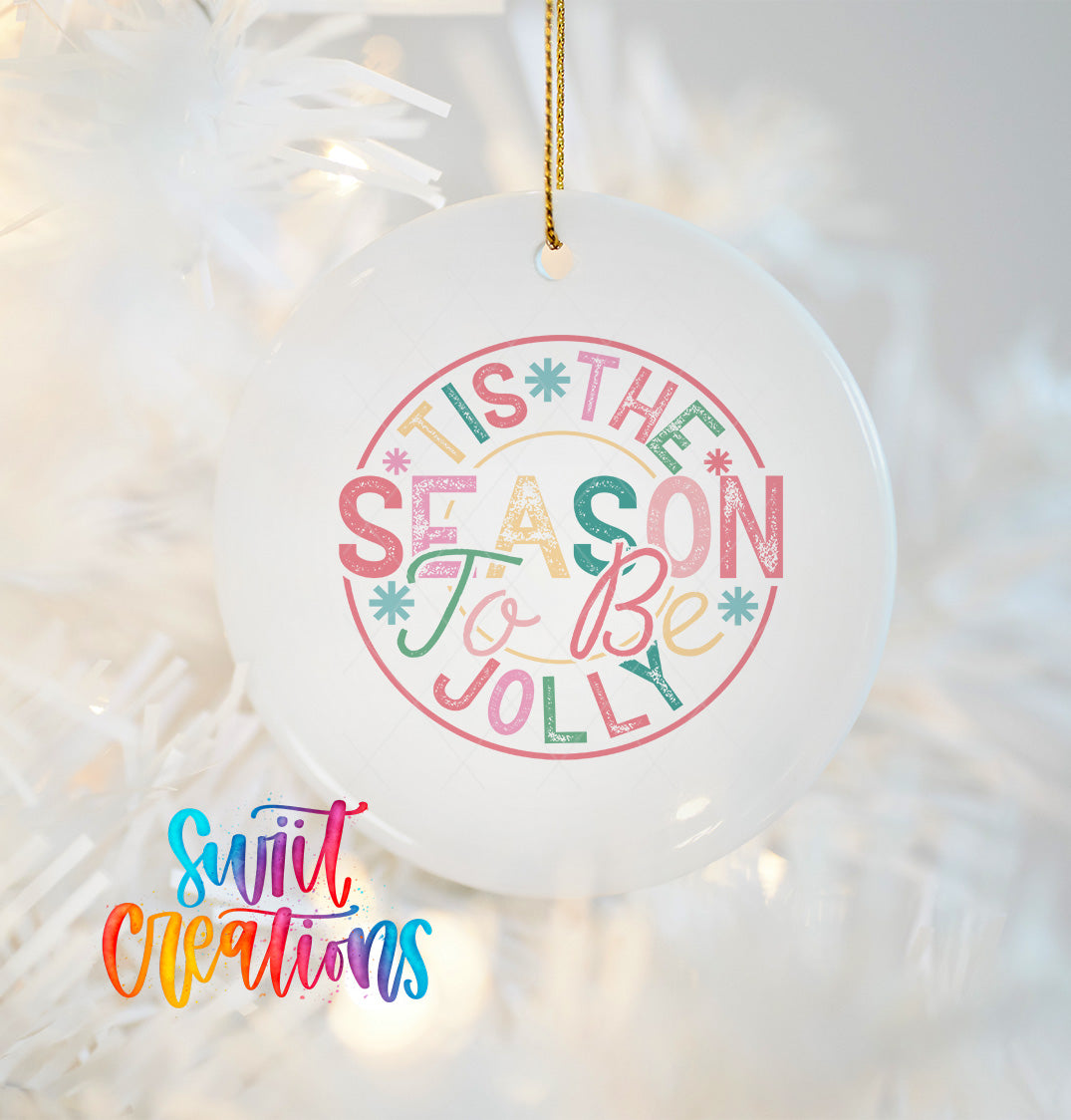 A white ceramic ornament with a colorful text design that says "TIS THE SEASON To BE JOYLY" hanging on a Christmas tree.