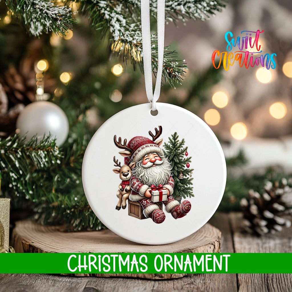 A white Christmas ornament featuring a Santa Claus figure holding a reindeer and a present, hanging on a tree branch with a blurred background of lights and greenery.