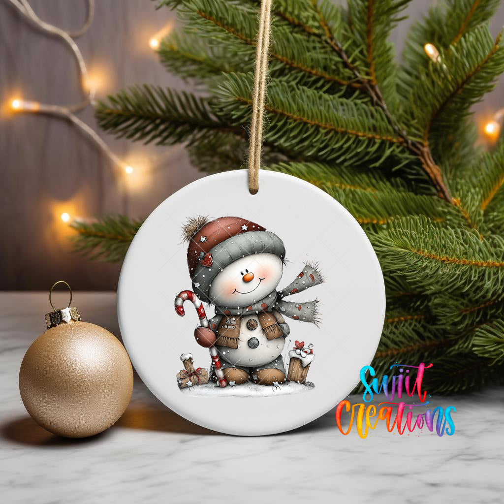 A white ceramic ornament featuring a snowman is displayed on a table, with a gold ornament and a Christmas tree in the background.