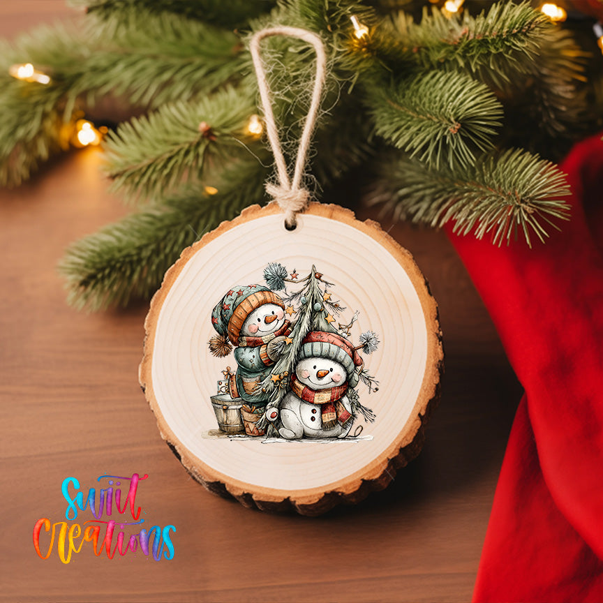 A wooden ornament featuring two snowmen is hanging on a Christmas tree, surrounded by pine branches and twinkling lights.