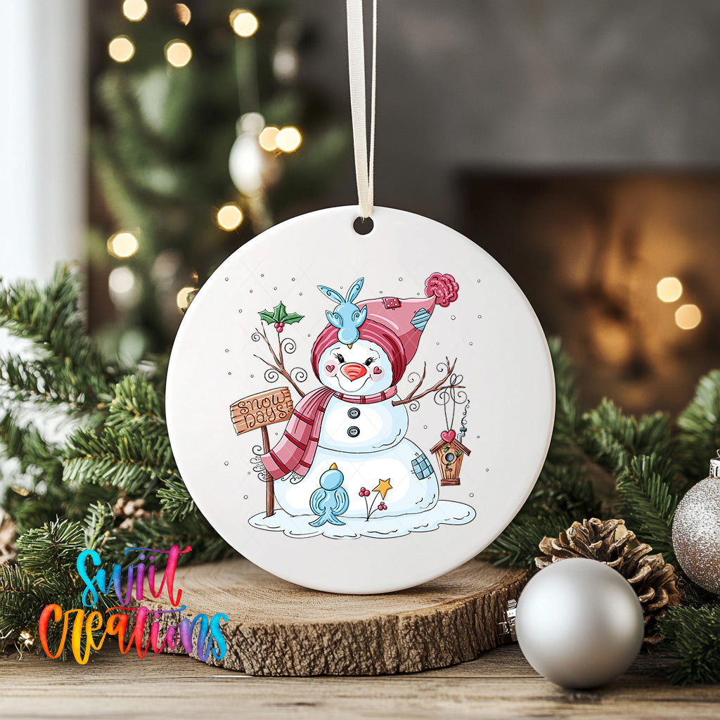 A white ceramic ornament featuring a snowman with a sign that says "Snowy Signs" is displayed on a wooden surface, surrounded by Christmas decorations and greenery.