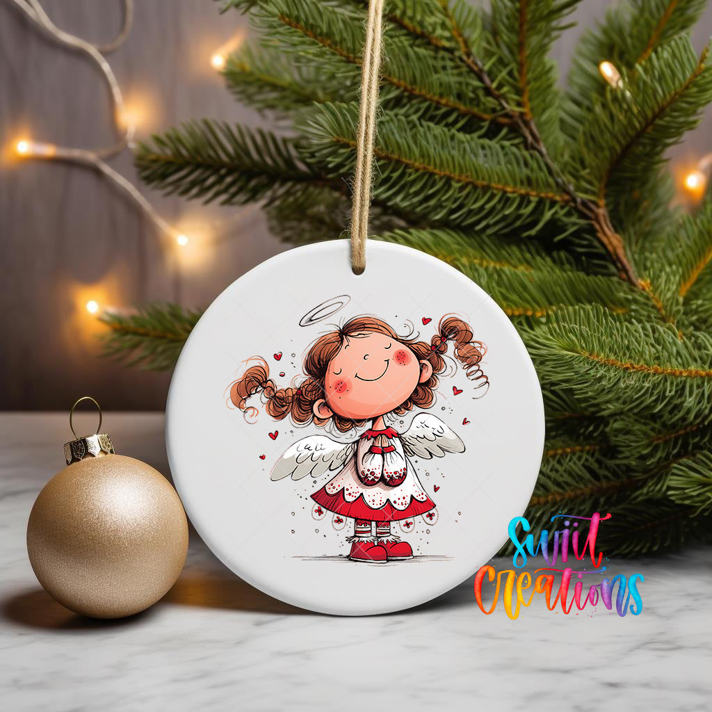 A white ceramic ornament featuring a smiling cartoon girl with curly hair and wings, surrounded by hearts, is displayed on a white marble surface next to a gold ornament and a Christmas tree.