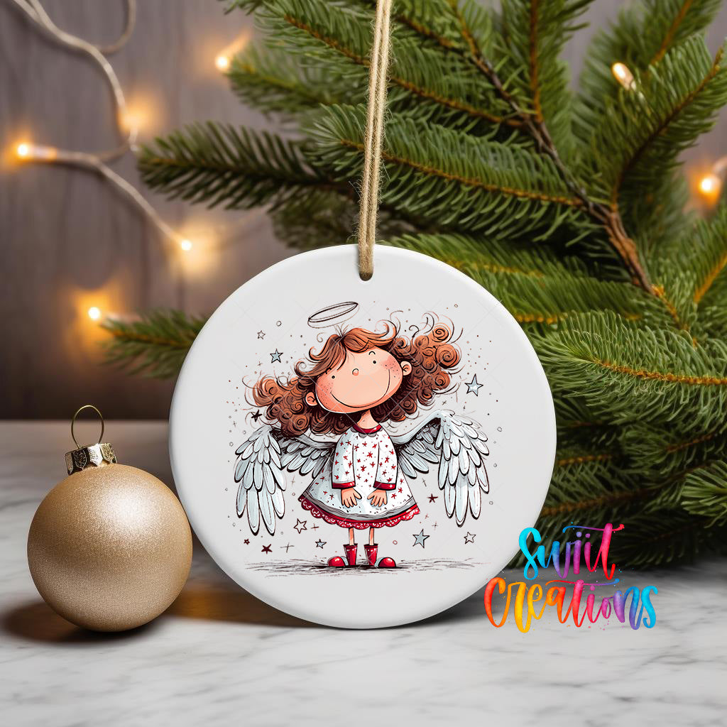 A white ceramic ornament featuring a smiling cartoon girl with curly hair and wings, surrounded by stars, is displayed on a table next to a gold ornament and a Christmas tree.