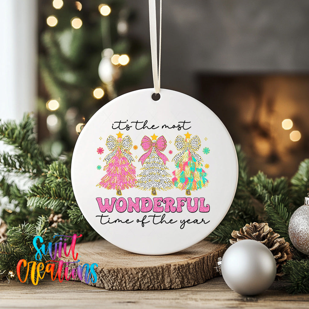 A white ceramic ornament with a festive design featuring three colorful Christmas trees and the text "It's the most wonderful time of the year" is displayed on a wooden surface, surrounded by holiday decorations such as ornaments and pine cones.