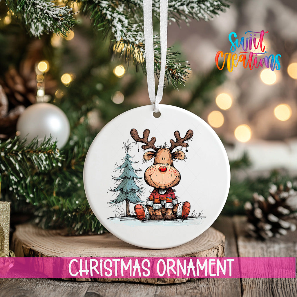 A white Christmas ornament featuring a cartoon reindeer sitting on a pine tree, with a blurred background of a decorated Christmas tree and twinkling lights.