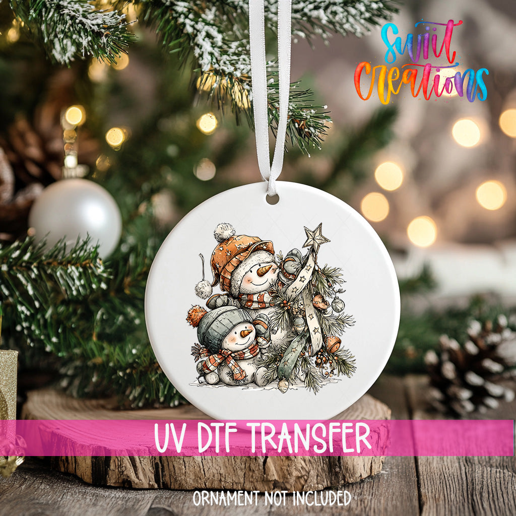 A white ornament with a snowman and star on a Christmas tree, with a pink ribbon around it.