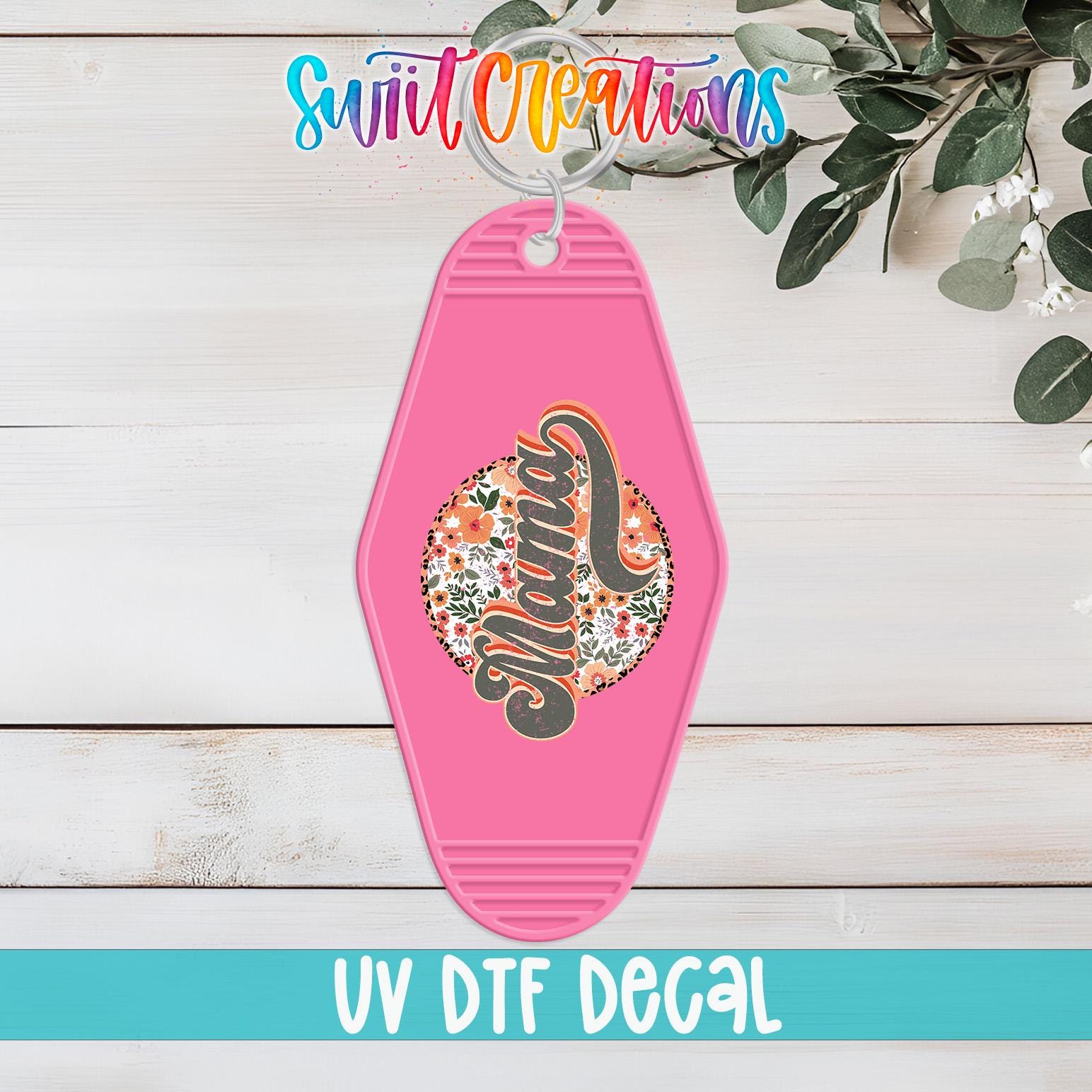 A pink keychain with a floral design and the word "Mama" on it, hanging on a wooden surface with a plant in the background.