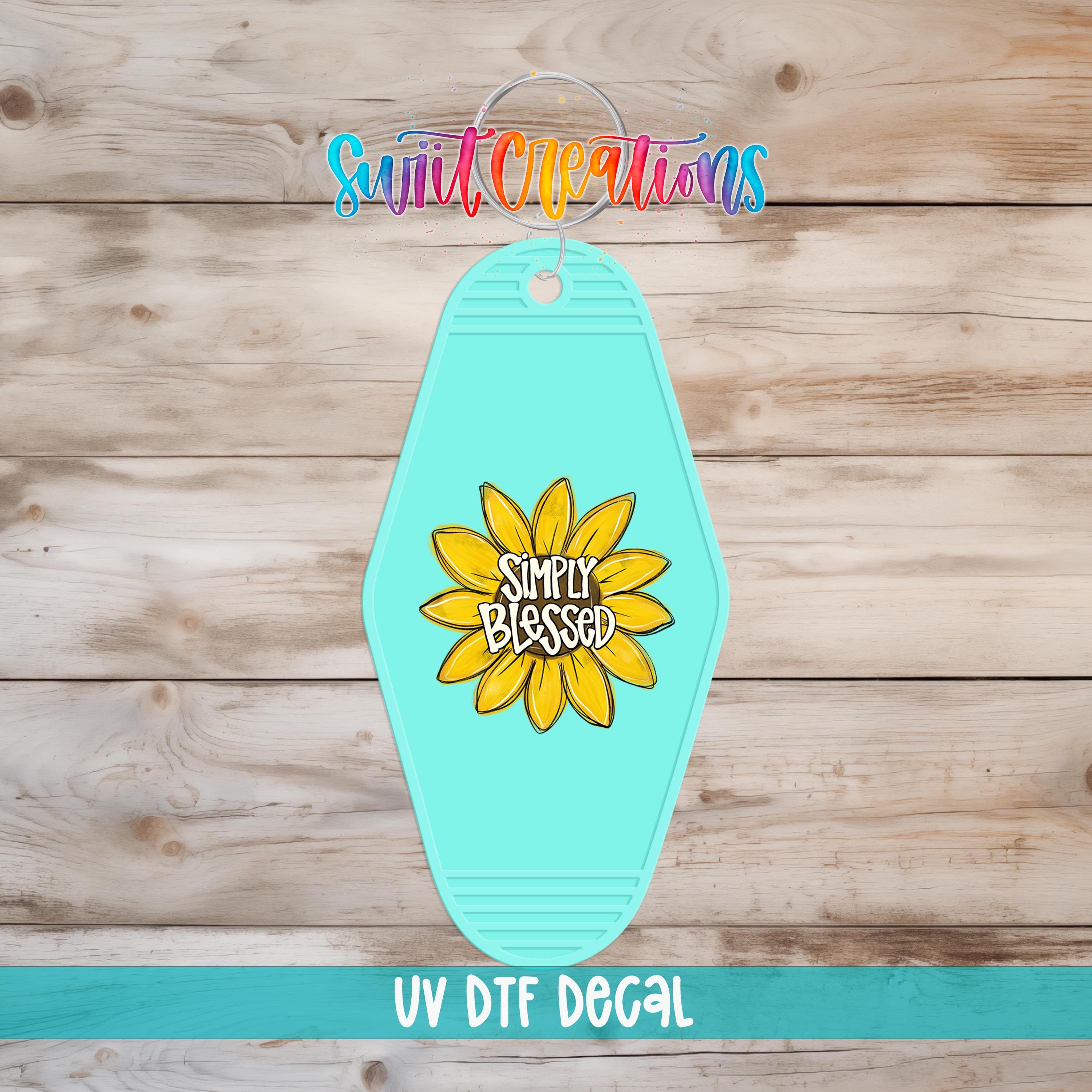 A turquoise keychain with a yellow sunflower design and the text "Simply Blessed" printed on it, placed on a wooden surface.