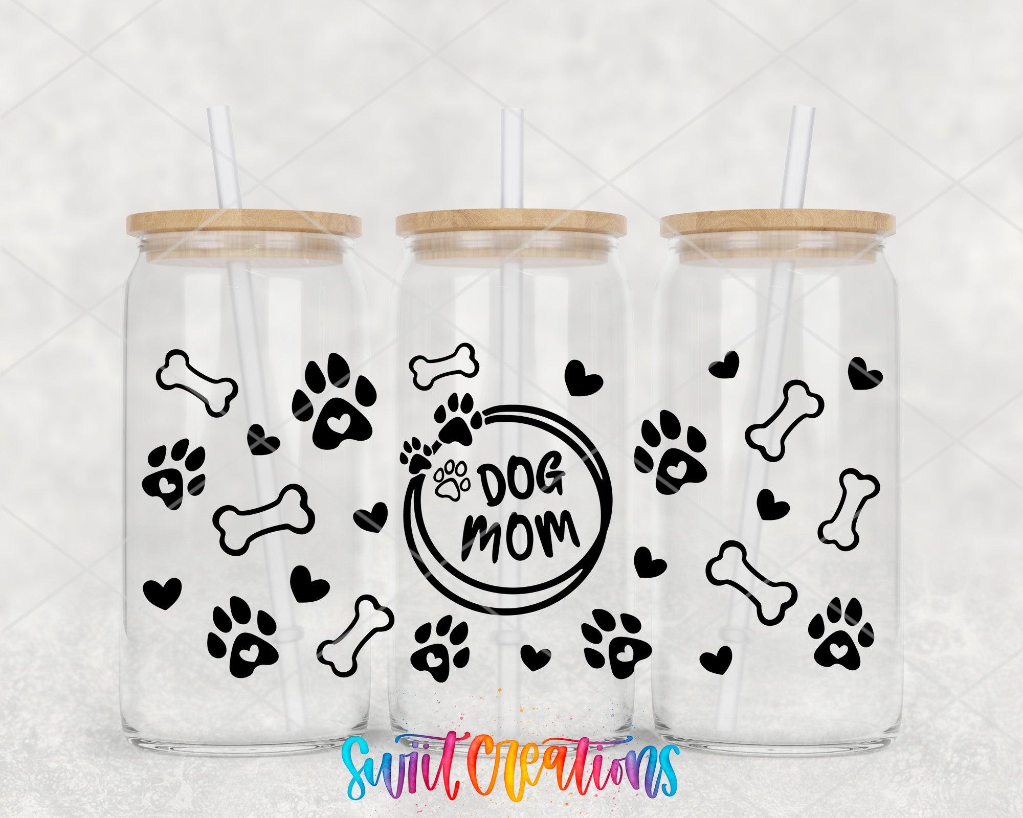 Three clear glass tumblers with wooden lids, each decorated with black paw prints and bones, and the phrase "dog mom" printed on them.
