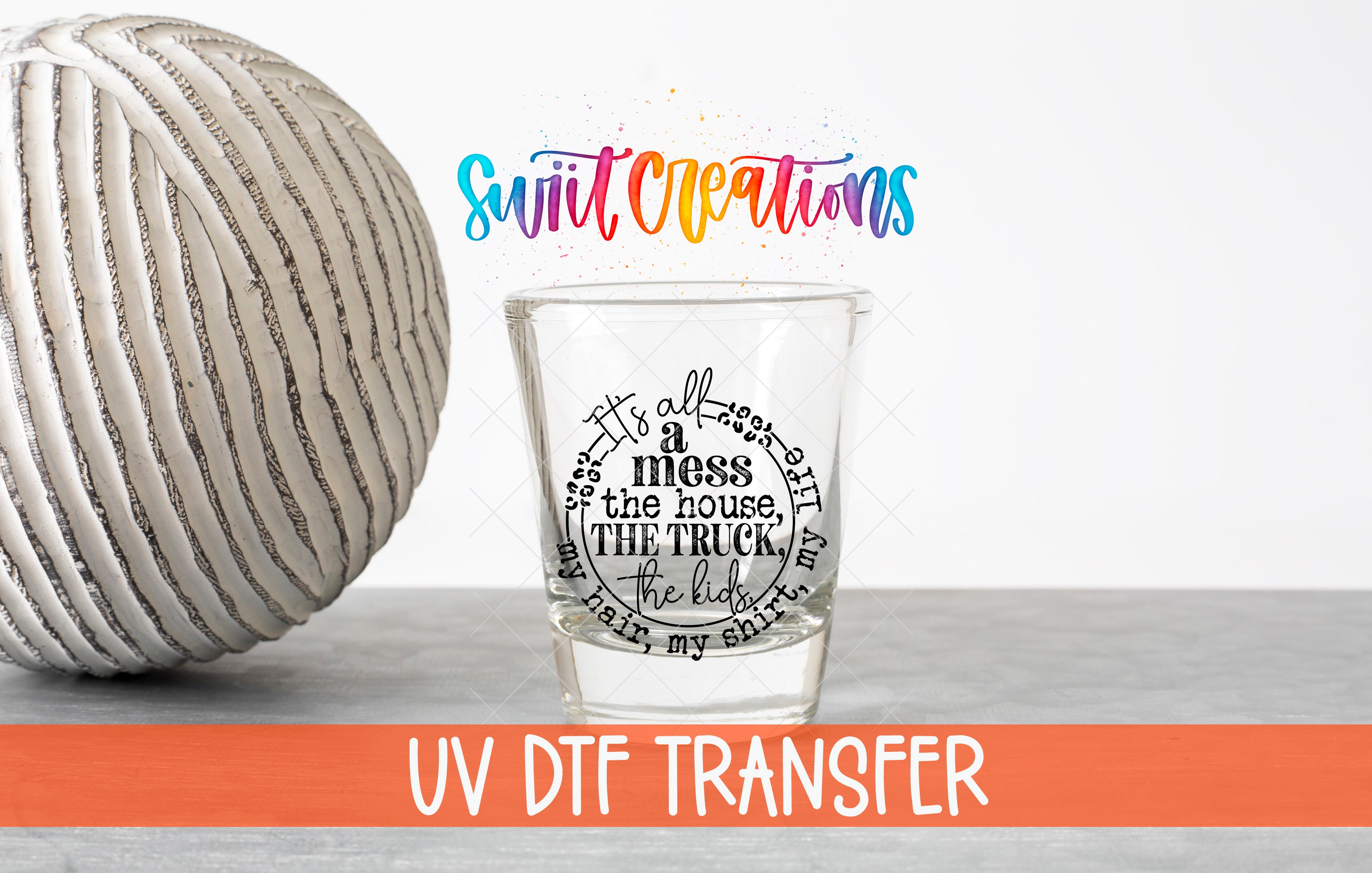 a shot glass with a message on it, placed next to a white ball on a gray surface. The shot glass has a message that reads "It's all a mess the house, the truck, the kids" in a circular design. The text "UV DFT TRANSFER" is also visible in the image.
