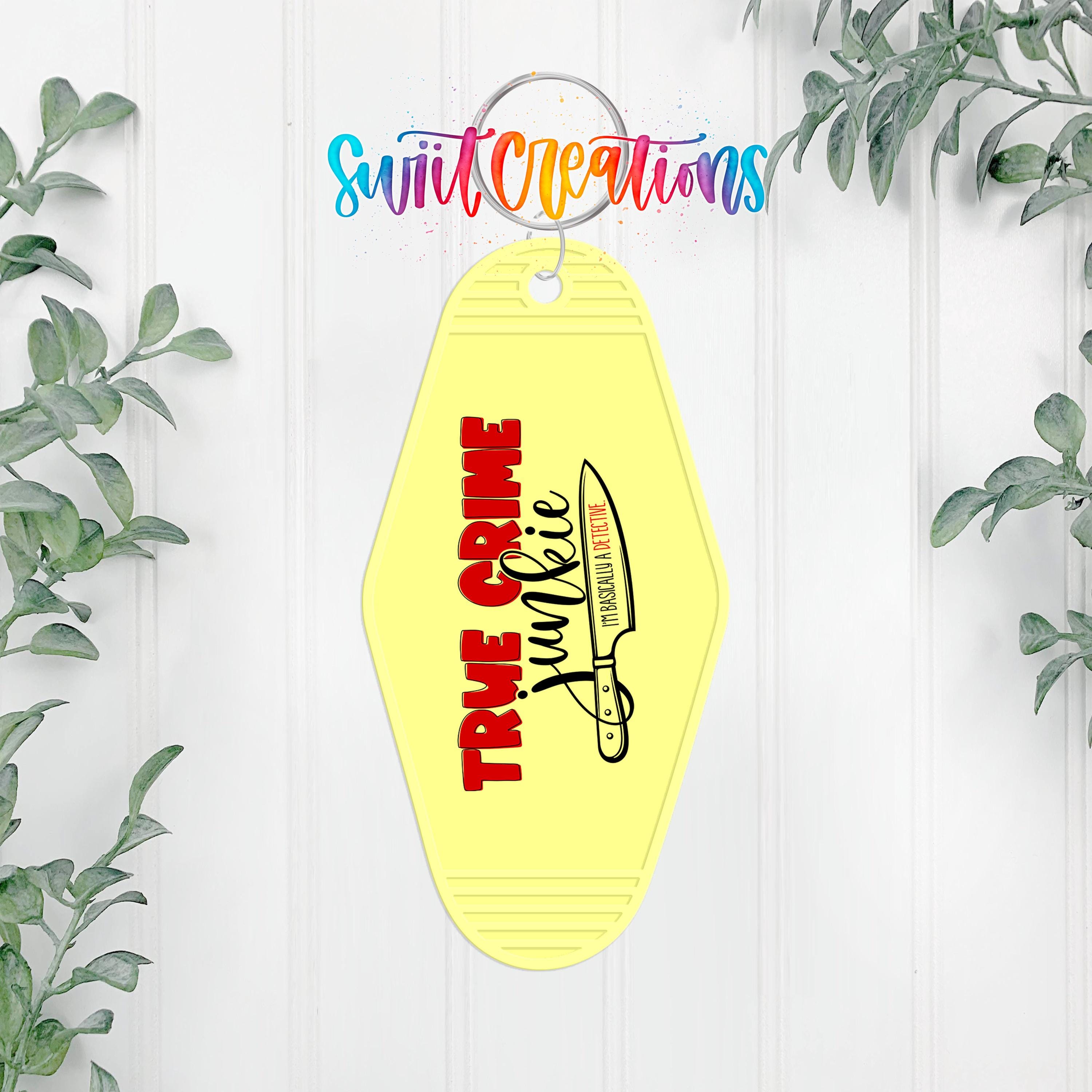 A yellow keychain with the text "TRUE CRIME" and a graphic of a knife, hanging on a white wall with greenery in the background.