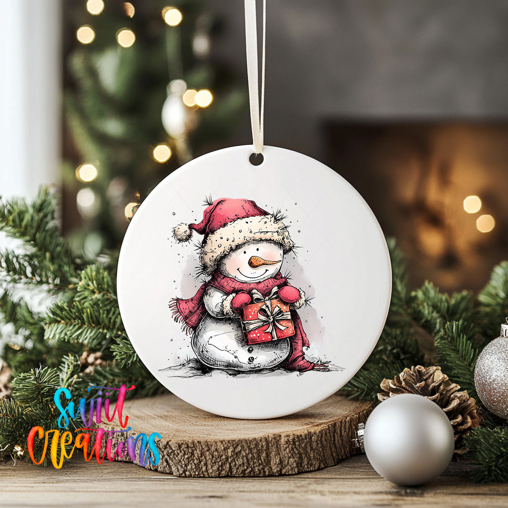 A white ceramic ornament featuring a snowman holding a gift, surrounded by Christmas decorations such as pine cones and ornaments, with a blurred background of a Christmas tree.