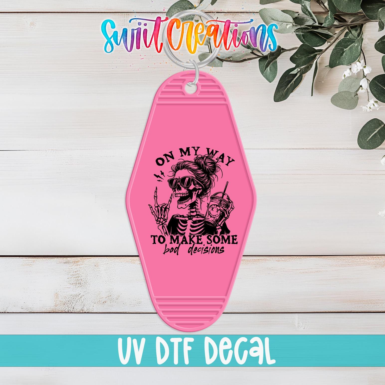 A pink keychain with a skull design and text, placed on a wooden surface with some greenery in the background.