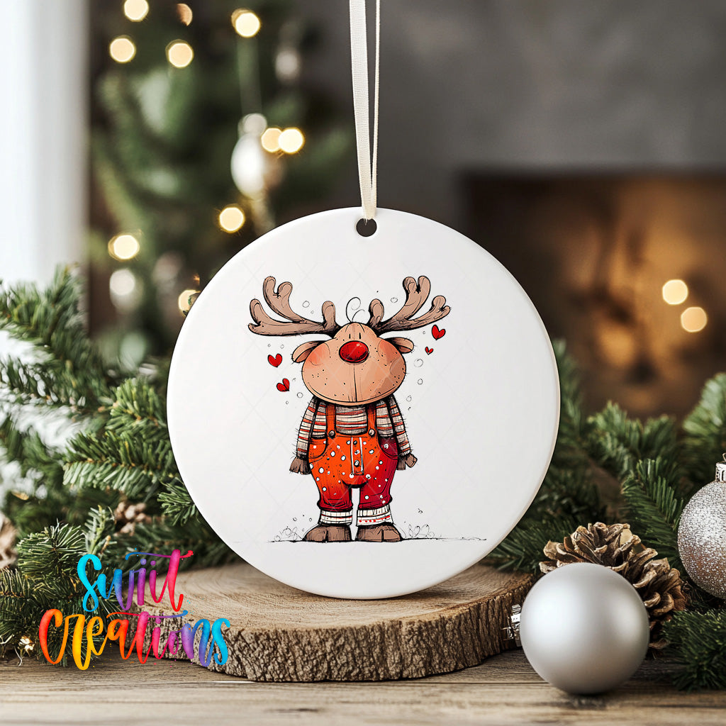 A white ceramic ornament featuring a cartoon reindeer with antlers and a red nose, surrounded by Christmas decorations such as pine cones and ornaments.
