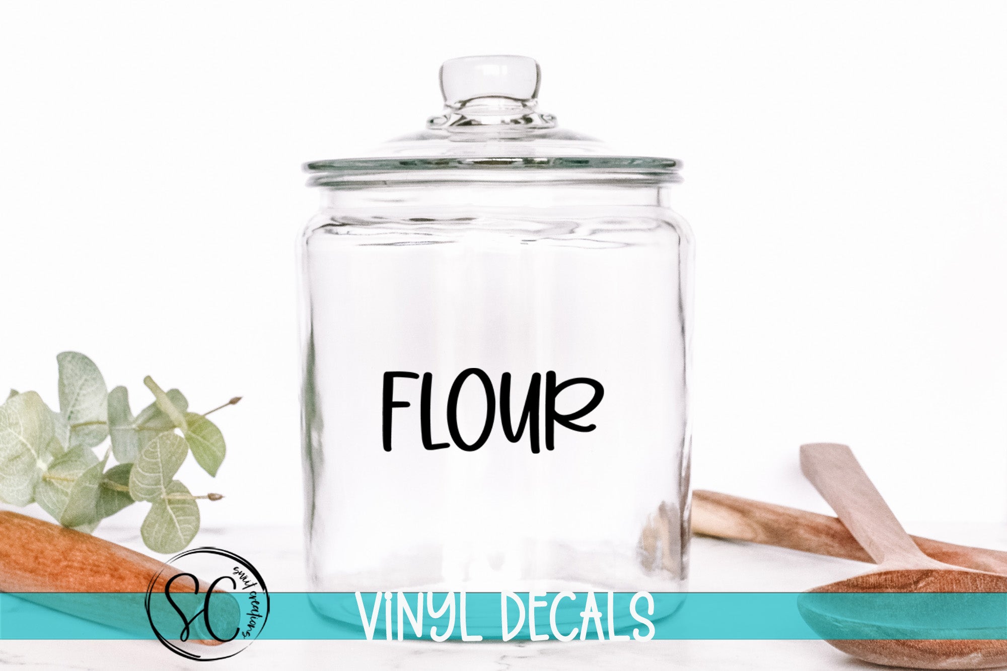 A clear glass jar with the word "FLOUR" printed on it, placed on a white surface next to some wooden utensils.