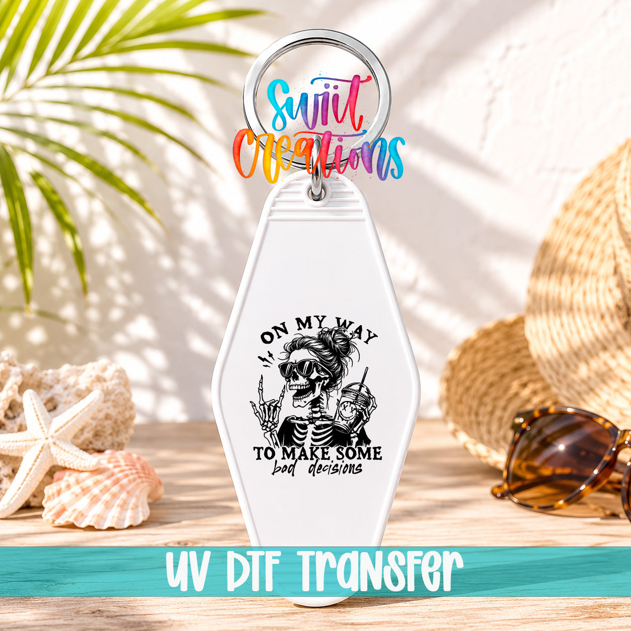 A keychain with a skeleton graphic and the text "ON MY WAY TO MAKE SOME BAD DECISIONS" printed on it, along with a colorful ring and sunglasses. The keychain is placed on a wooden surface, with a straw hat and sunglasses visible in the background.