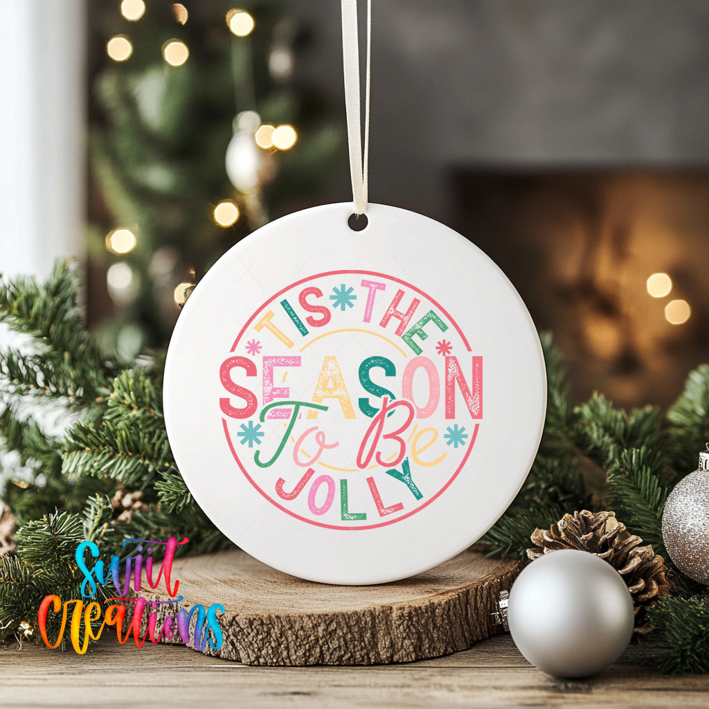 A white ceramic ornament with a colorful text design that says "THIS SEASON TO BE JOYOUS" is displayed on a wooden surface, surrounded by Christmas decorations such as pine cones and ornaments.