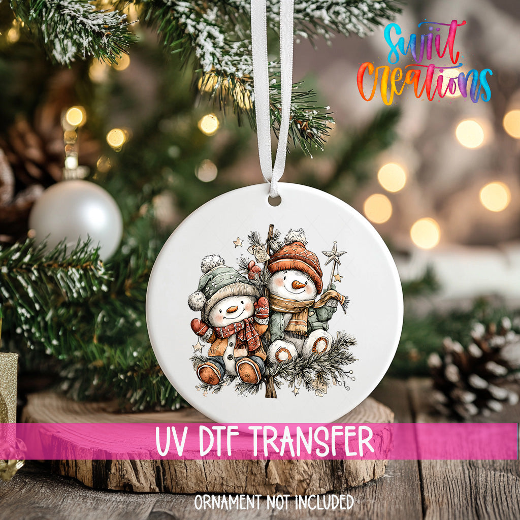 A white ornament with a snowman and star on a Christmas tree, with a pink ribbon around it.